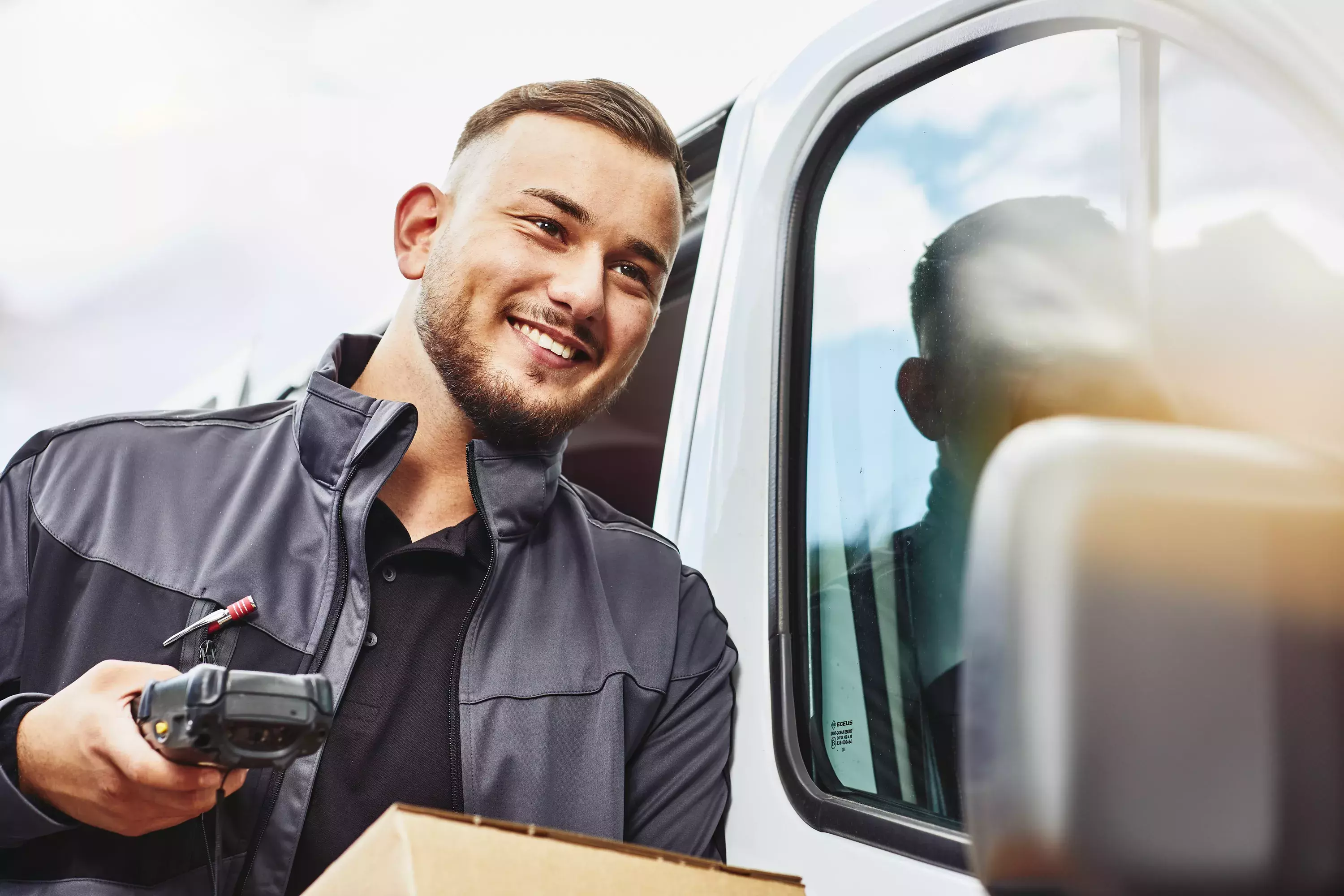 Close up - Smiling male holding a scanner and package standing next to a delivery van.
