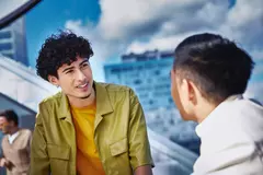 Male in yellow shirt and male, standing outside on a balcony, talking